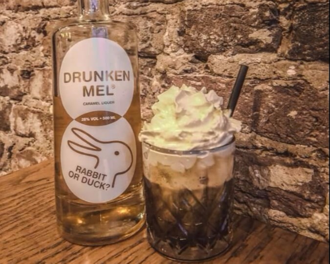 Fles Drunken Mel tegen de muur met een glazen kop koffie met slagroom